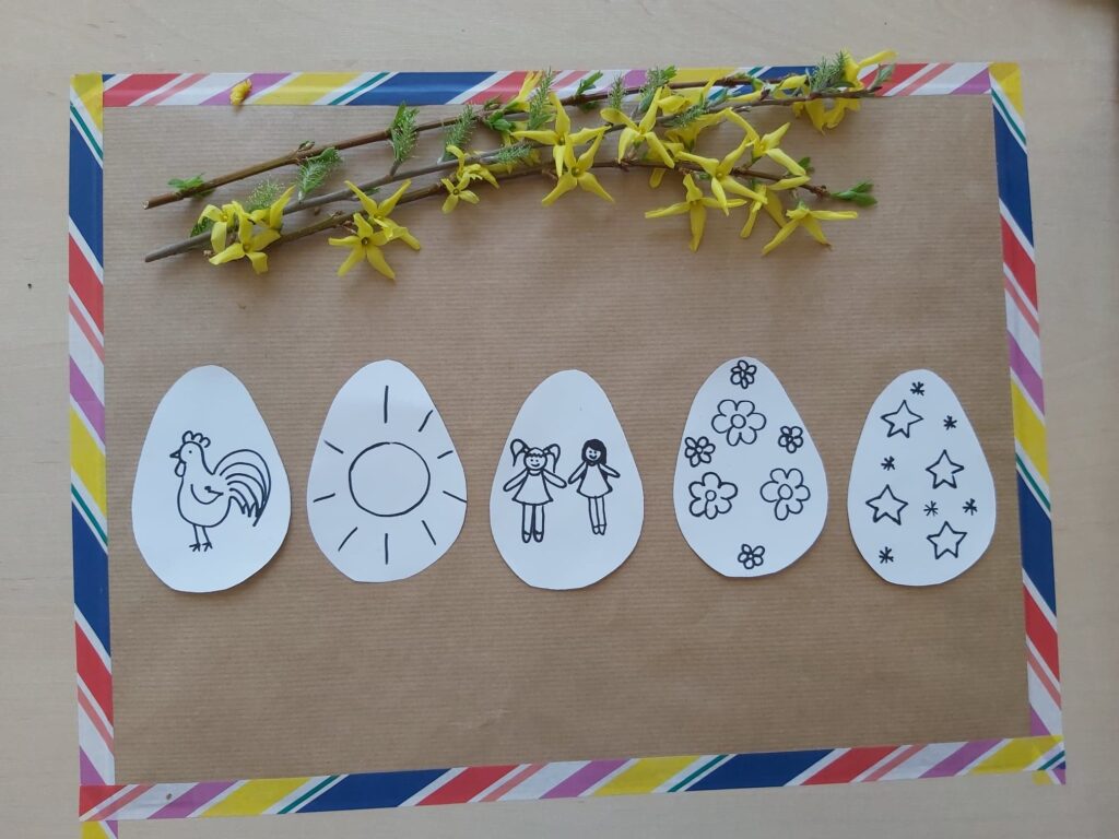 Na kartce leżą papierowe jajka z rysunkami koguta, słońca, laleczek, kwiatków, gwiazdek