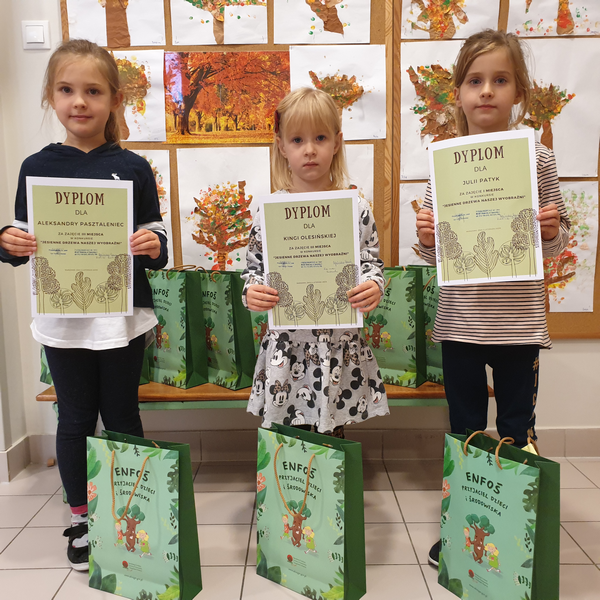 Trzy dziewczynki stoją na tle rysunków drzew, trzymają w rękach dyplomy, przed nimi stoją papierowe, zielone torebki.