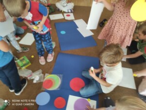 Grupa dzieci na dywanie przykleja kropki różnej wielkości do kartonu. 