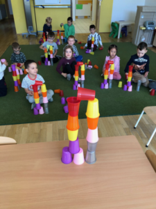 Na pierwszym planie podwójna wieża z kolorowych kubków, w tle grupa dzieci układa taką samą wieżę