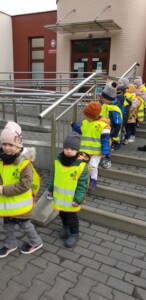 Dzieci w kamizelkach odblaskowych schodzą za schodów przy budynku Przedszkola.